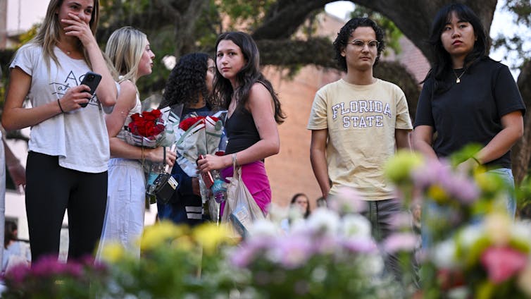 A group of young people, some holding flowers, stand in front of a collection of flowers on the ground
